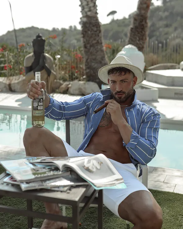 Maximo Garcia de chapéu e camisa listrada, relaxando à beira da piscina com revistas na mesa